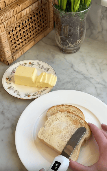 A hand spreads butter onto a slice of toasted bread with a knife, next to a floral plate holding a block of butter on a marble countertop. A potted plant and wicker box are in the background.