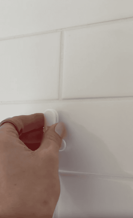 A hand with a gold ring presses a small white adhesive hook onto a white tiled wall, perfect for organizing essentials in your shower or creating order in your home sauna retreat.