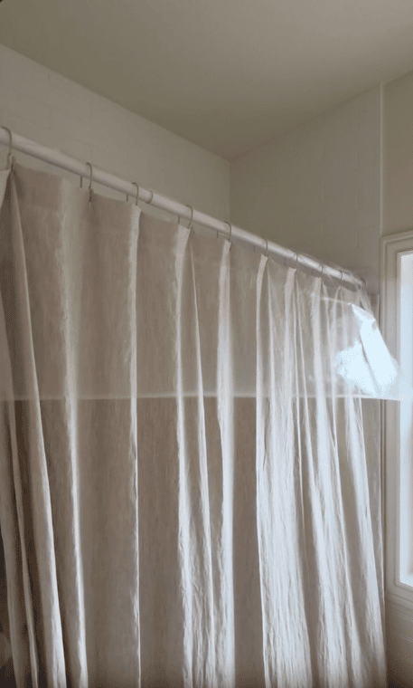 A beige shower curtain hangs from a white shower rod in a bathroom with white tiled walls and a frosted window to the right, creating a peaceful shower sauna vibe for your own relaxing retreat.