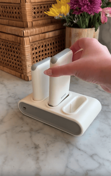A hand places a white, rectangular electronic device into a matching charging dock on a marble countertop. Wicker baskets and a vase of colorful flowers are in the background.