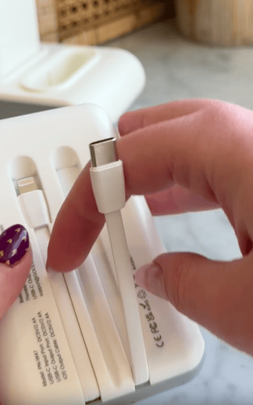A person with purple, dotted nail polish holds a white USB-C charging cable from a white charging station on a marble countertop.