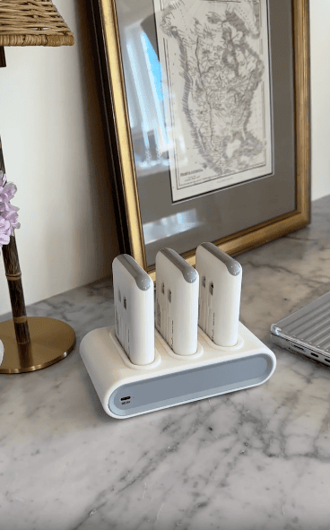 Three white handheld devices are charging vertically in a white charging dock on a marble countertop. Behind them is a framed map, and part of a tablet or keyboard is visible on the right.