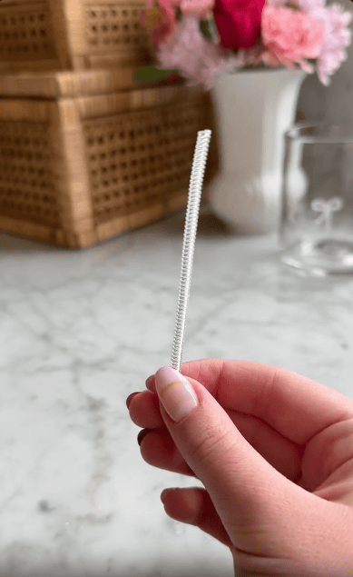 A hand holding a short white pipe cleaner in front of a marble countertop, with a mini waffle maker, a wicker basket, a glass jar, and pink flowers blurred in the background.