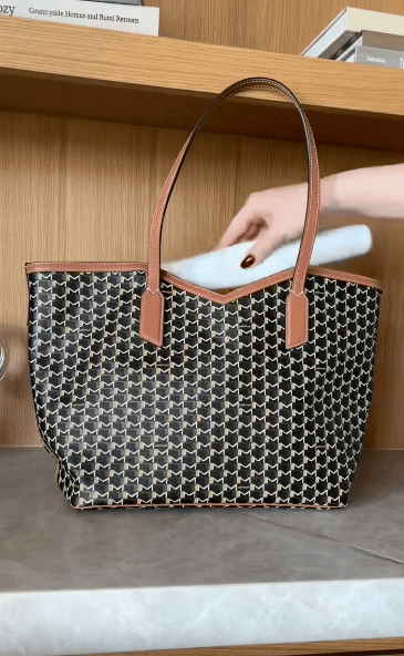A person places a rolled-up white towel into a patterned tote bag with brown handles, set on a gray surface in front of wooden shelves.