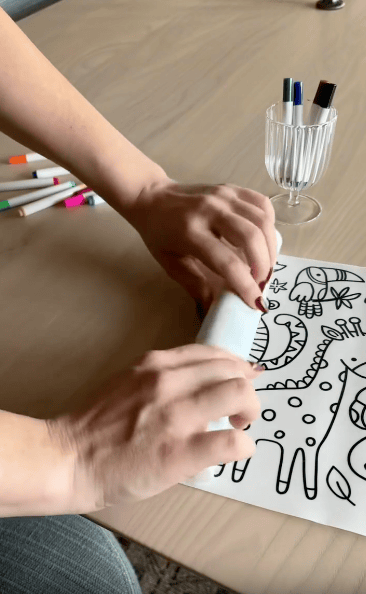 A person rolls up a sheet of black-and-white animal drawings on a table, with colored markers and a glass holding pens nearby.