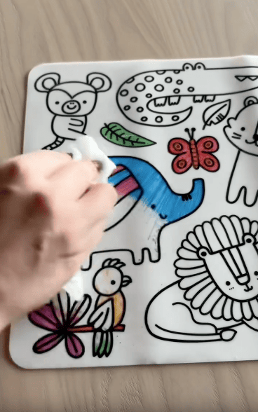 A hand wipes blue marker off a drawing of an elephant on a reusable coloring board featuring outlined animals, including a lion, crocodile, monkey, butterfly, and bird.