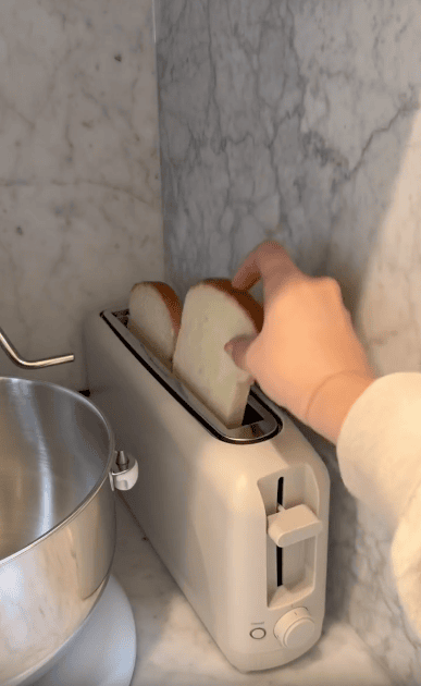 A hand is placing two slices of bread into a white toaster on a marble kitchen countertop next to a metal mixing bowl and a mini waffle maker.