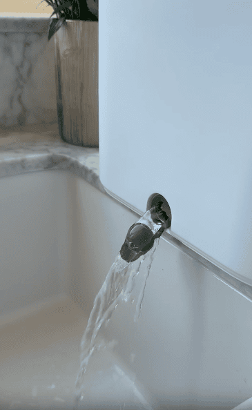 Water flows out of a modern metal spout attached to the side of a white bathtub, with a marble countertop and a wooden planter visible in the background.