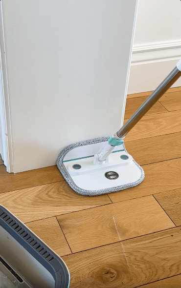 A flat mop with a grey and white microfiber pad is being used to clean the wooden floor along the base of a white wall.