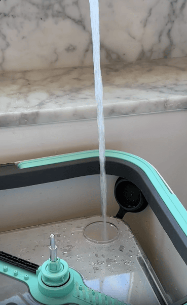 A stream of water flows from a faucet into a rectangular container with a mop and cleaning tools inside, set against a marble countertop background.