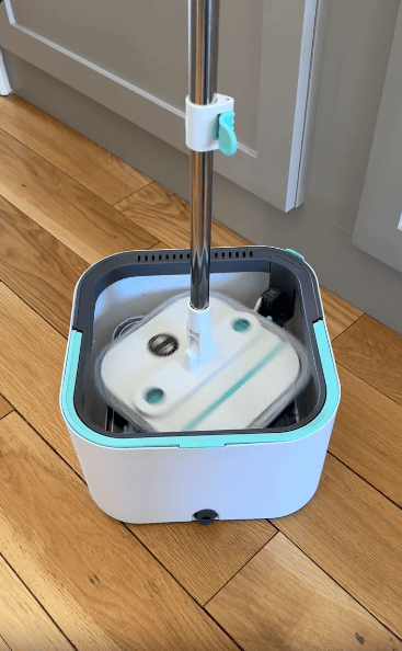 A flat mop is being rinsed or spun inside a square, white and teal self-cleaning mop bucket on a wooden floor.