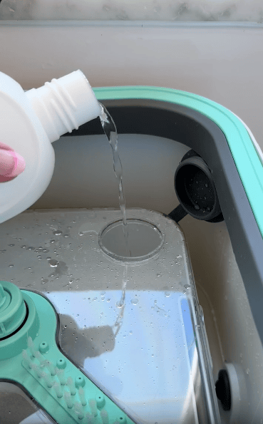 A hand pours liquid detergent from a white bottle into the designated compartment of a light green and gray cleaning bucket or mop system, with some water and cleaning brushes visible.