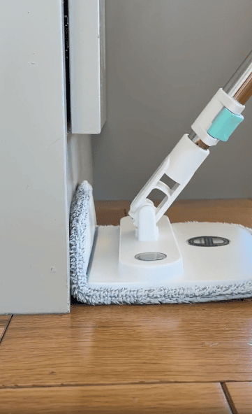 A flat microfiber mop cleans a narrow gap between a wall and a piece of furniture on a wooden floor.