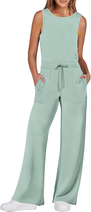 Woman wearing a sleeveless, light sage green jumpsuit with wide-leg pants, patch pockets, a drawstring waist, and white sneakers. She has her hands in her pockets and is smiling.