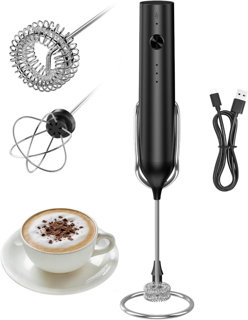 A black handheld milk frother with two detachable whisk attachments, a USB charging cable, and a cup of cappuccino with foam and chocolate shavings on a saucer, shown on a white background.
