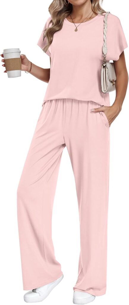 A woman wearing a light pink short-sleeve lounge set with wide-leg pants, white sneakers, and a crossbody bag holds a coffee cup. Her wavy hair is down, one hand in her pocket—ready to take on Friday Deals in effortless style.