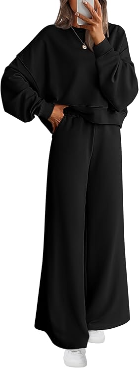 A person wearing a loose-fitting black sweatshirt and matching wide-leg pants poses while holding a phone. They have long brown hair and are also wearing white sneakers and a necklace.