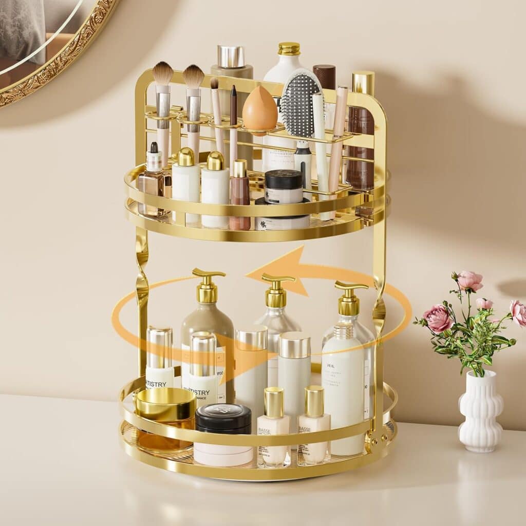 A gold two-tier rotating organizer holds various skincare, makeup, and hair products on a white surface. There are brushes, bottles, and jars, with a small vase of pink flowers and a mirror partially visible in the background.