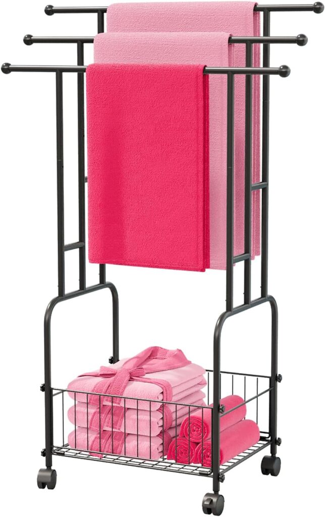 A black metal towel rack on wheels holds two pink towels and one light pink towel draped over bars, with folded pink towels and a pink bathrobe stored in a wire basket at the bottom.