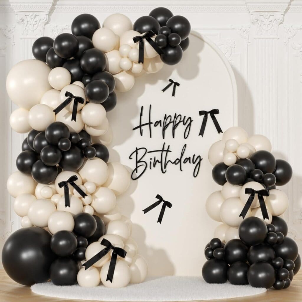 A decorative black and white balloon arch with black bow accents surrounds a "Happy Birthday" sign on a white ornate wall background.