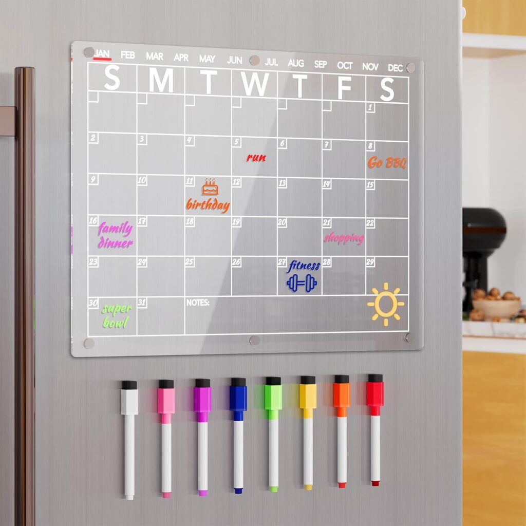 A clear acrylic monthly calendar, part of our Friday Deals, is attached to a fridge with colorful events written on it. Below are eight vibrant dry-erase markers lined up neatly. Kitchen shelves and appliances can be seen in the background.