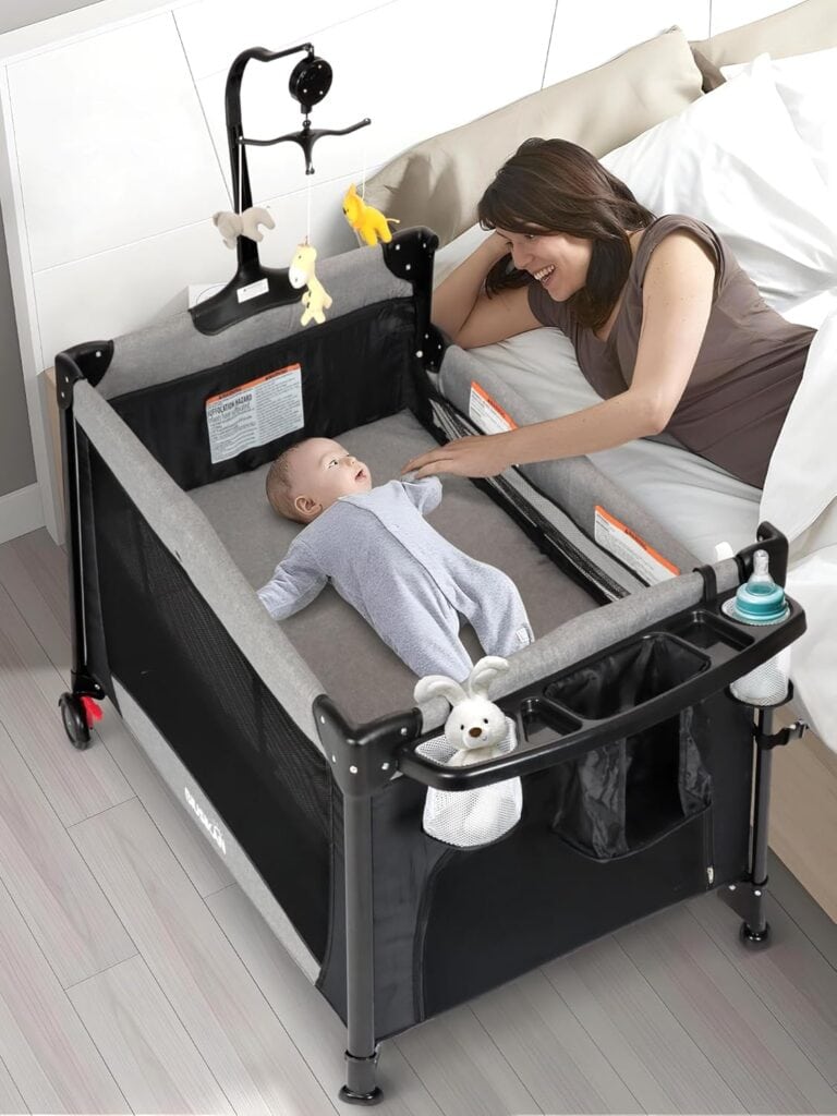 A woman lies in bed smiling at a baby who is lying in a gray and black Pack and Play bassinet beside the bed. The playpen has hanging toys and baby items attached, and the baby looks up at the woman. Get 50% off for a limited time!.