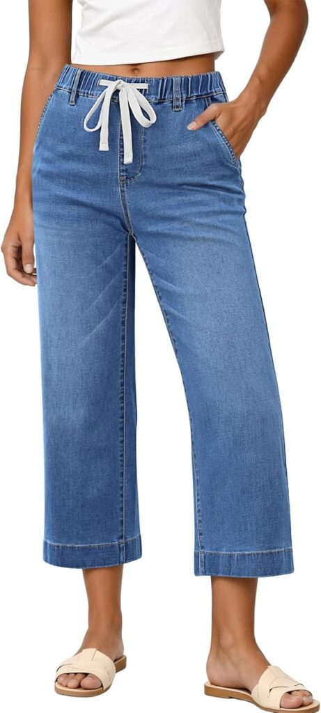 A person wearing cropped blue denim pants with a white drawstring, a white sleeveless top, and beige sandals, with one hand in a pocket.