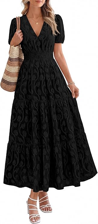 A woman wearing a black, short-sleeve, V-neck, ankle-length dress with a subtle paisley pattern, holding a straw tote bag and wearing white strappy sandals.