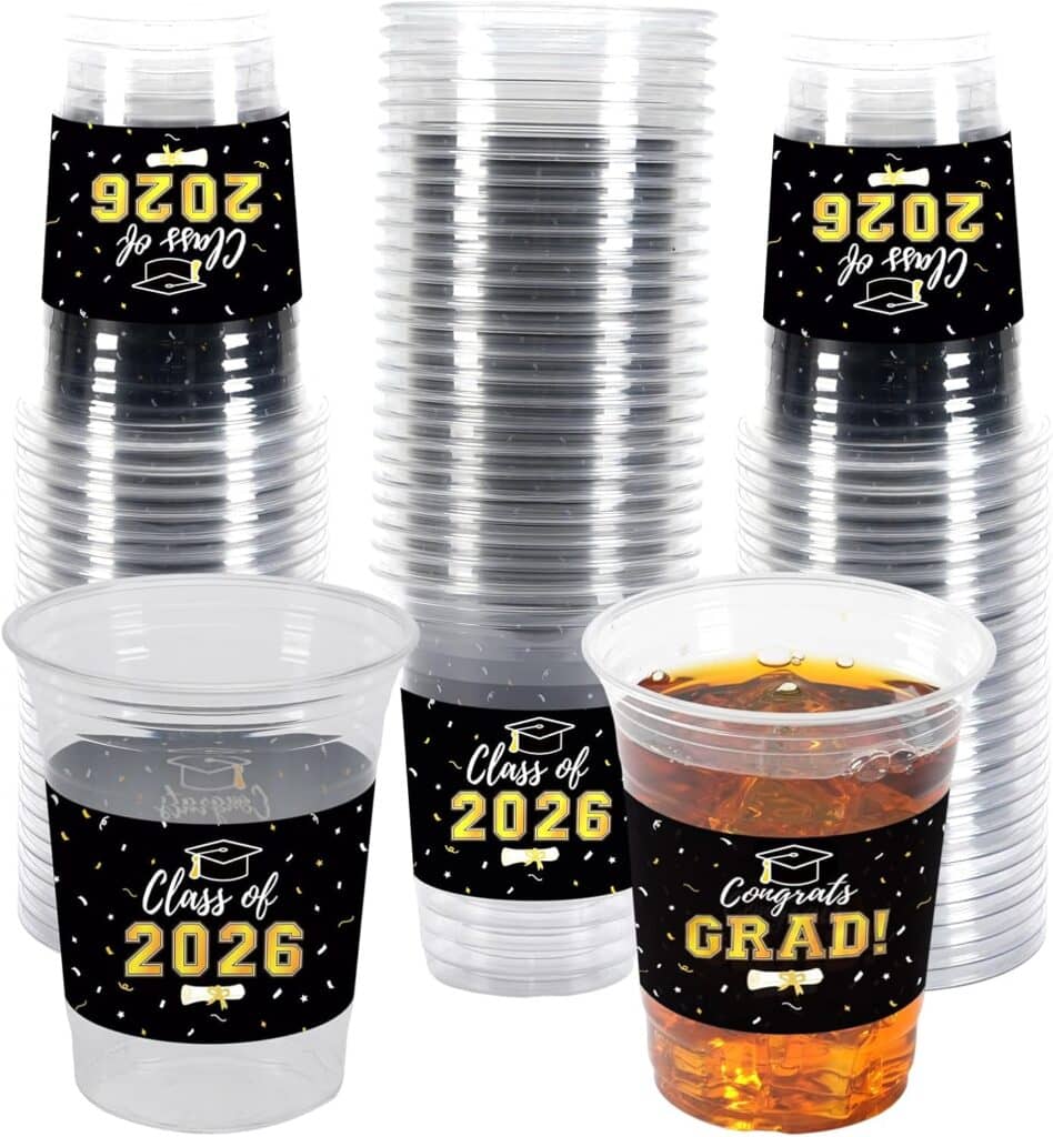 Stacks of clear plastic cups with black and gold labels saying “Class of 2026” and “Congrats Grad!”—perfect for your celebration. One cup is filled with an orange drink, making these a must-have for Friday Deals.