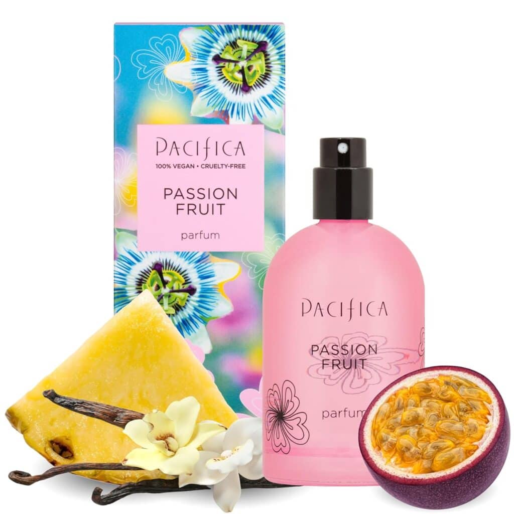 A pink bottle of Pacifica Passion Fruit perfume is placed in front of a colorful box with passion fruit flowers. Slices of pineapple, passion fruit, vanilla bean, and a white flower are arranged beside the bottle.