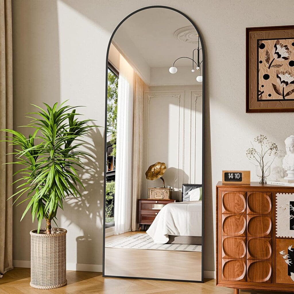 A large arched mirror reflects a bright bedroom with a white bed, vintage decor, and a window. A potted plant and a wooden cabinet with a digital clock and art pieces sit beside the mirror on a light-colored wall.