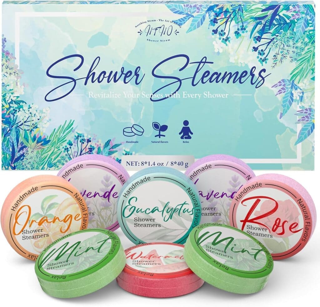 A box labeled "Shower Steamers" is displayed behind six colorful round shower steamer tablets in scents: Orange, Eucalyptus, Rose, Lavender, and Mint. The packaging features a floral, botanical design.