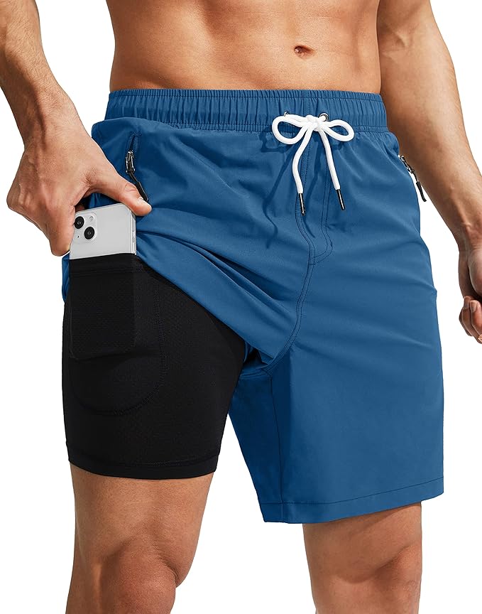 A person wearing blue athletic shorts with a white drawstring reveals black compression shorts underneath, which have a side pocket holding a smartphone.