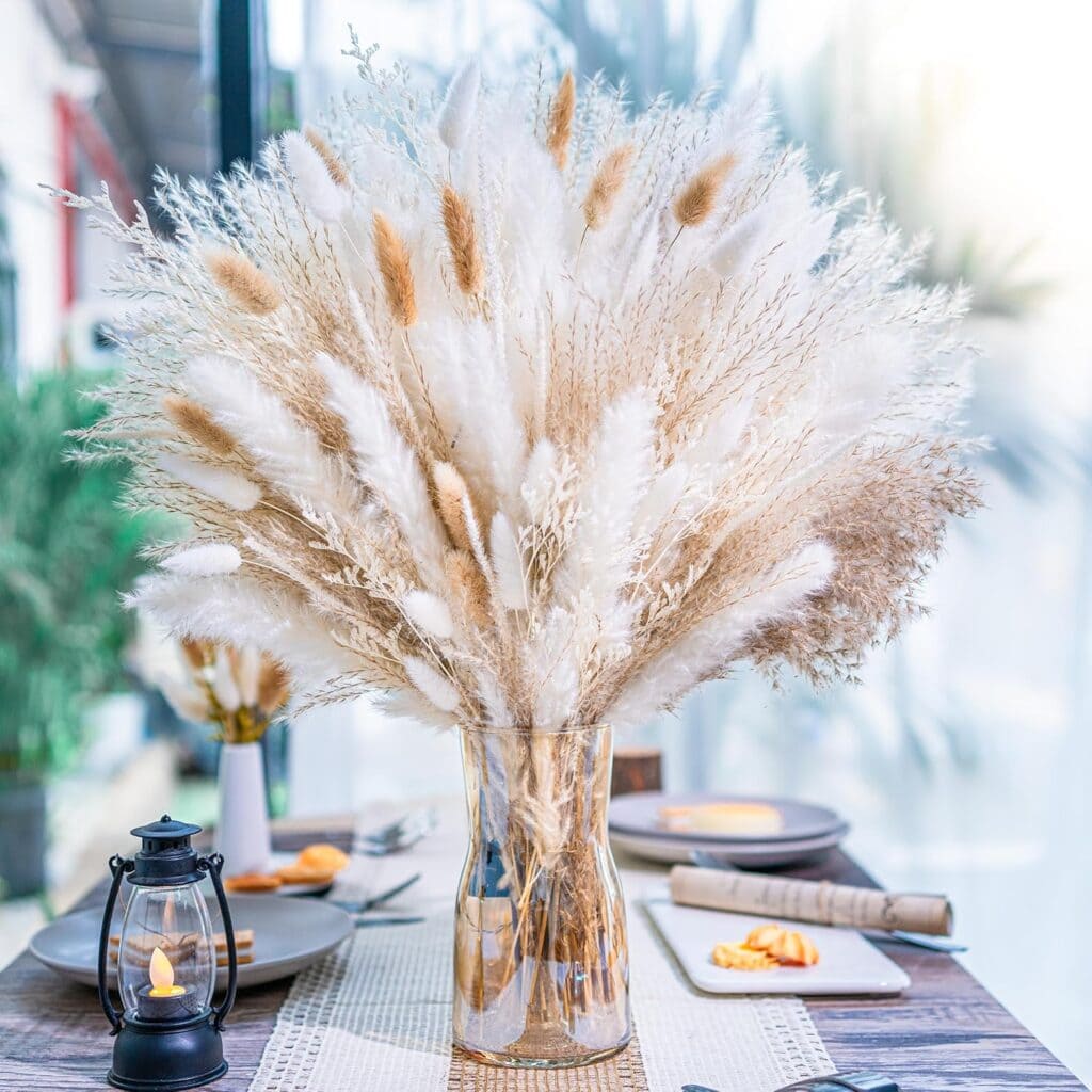 A glass vase filled with a large decor bouquet of natural pampas grass sits on a set dining table, decorated with plates, cutlery, and a small lantern in a bright, modern room.