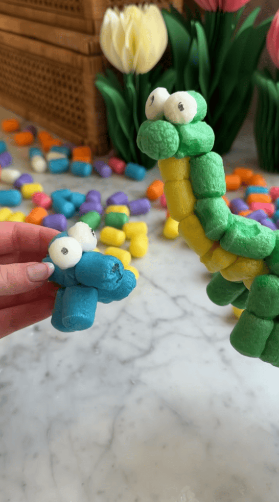 Two colorful caterpillars made from Magic Noodles with googly eyes are held up, surrounded by more packing peanuts and artificial tulips on a marble surface—a fun foam crafts project perfect for kids.