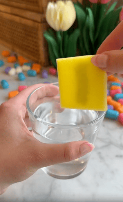 A hand holds a yellow sponge above a glass of water, with colorful magic noodles and tulips in the blurred background on a marble surface—perfect inspiration for kids’ foam crafts.