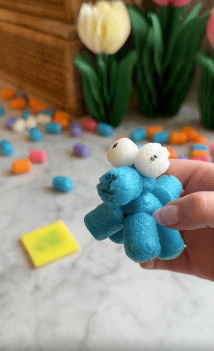 A hand holds a small, blue, bug-like figure made from colorful Magic Noodles with marker-drawn eyes and mouth. More packing peanuts and artificial plants are scattered in the blurred background—perfect for kids' foam crafts.