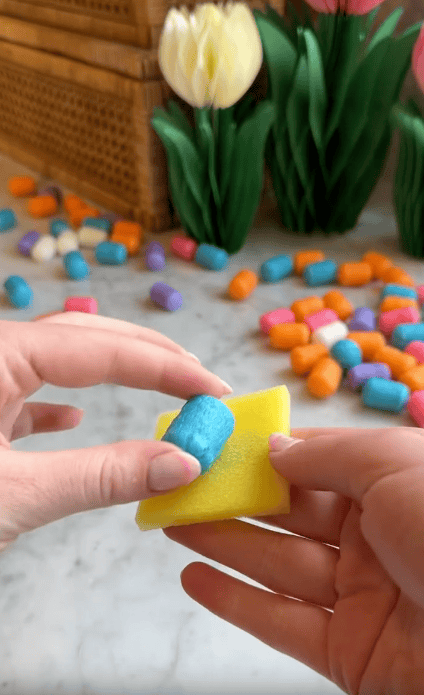 A person presses a blue Magic Noodle onto a small yellow sponge. Colorful craft peanuts are scattered on a marble surface, with artificial tulips and a wicker basket in the background—perfect for creative kids' foam crafts.