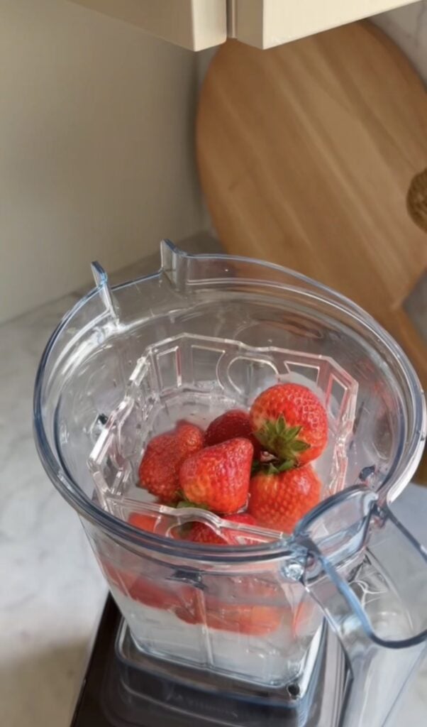A blender cup filled with whole strawberries, ice cubes, and water sits on a kitchen counter, perfect for a refreshing drink after bike training. A wooden cutting board is visible in the background.