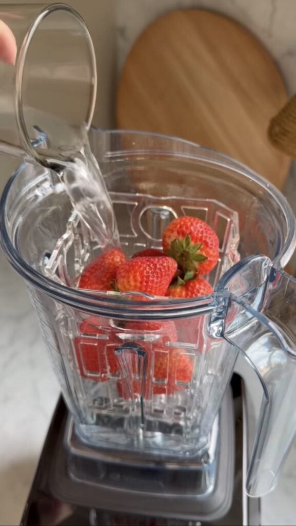 A hand pours water from a glass into a blender filled with fresh strawberries, set on a kitchen countertop after an intense bike training session.