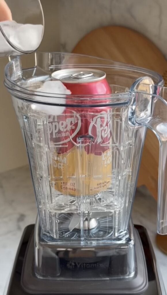 A hand pours ice cubes into a blender with a whole, unopened can of Dr Pepper soda on a kitchen counter—a true back saver for anyone needing quick refreshment after bike training. A cutting board sits in the background.