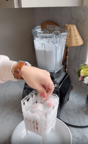 A hand holds a plastic basket with strawberries, covered in frothy white liquid, above a plate. In the background, beside a bike training handle and a blender with white liquid inside, sits a lamp and a vase of flowers on the marble countertop.
