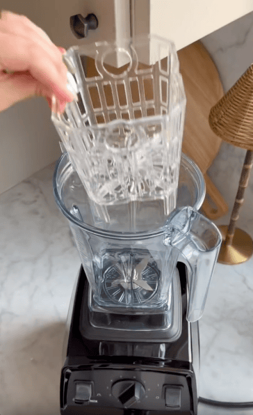 A person places a clear plastic dishwashing basket into a blender pitcher on a black blender base, set on a marble countertop next to a lamp, a wooden board, and a back saver for bike training.