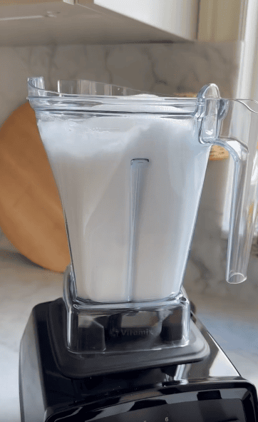 A blender filled with a white, frothy liquid is blending on a kitchen counter. Nearby, a back saver bike training handle rests against a wooden cutting board, with sunlight streaming in through the window in the background.