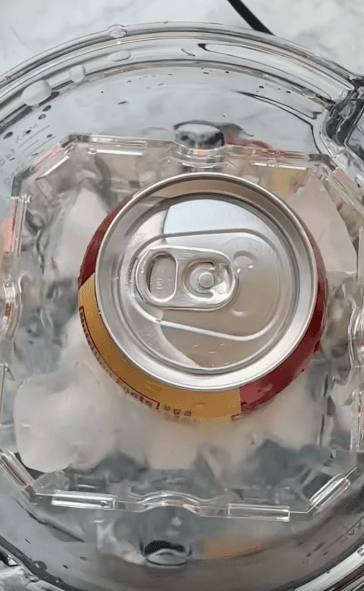 A top-down view of a blender filled with ice cubes and a closed soda can, showing the can placed upright among the ice inside the blender jug—perfectly chilled after an intense bike training session.