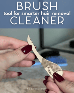 A person with dark red nails holds a small, gold brush cleaner tool with grooves and a textured handle—perfect as a back saver for smarter hair removal. A blue hairbrush, mirror, and even a bike training handle appear in the blurry background.