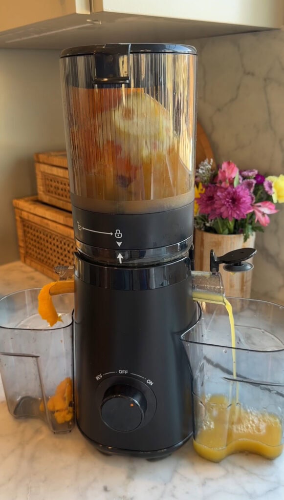 A black juicer is processing orange slices, with juice flowing into a clear container and pulp collected on the side. A bouquet of flowers and woven baskets add charm to the counter space on a marble countertop.