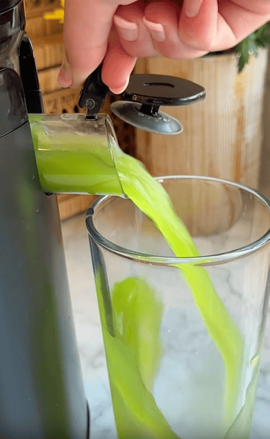A hand holds a small spout, pouring bright green juice from a juicer into a tall clear glass on a marble countertop, showcasing the perfect kitchen appliance for fresh drinks without taking up much counter space.