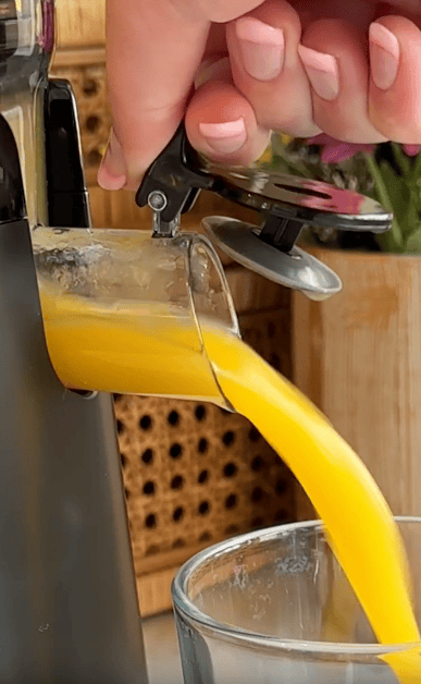 A close-up of a hand operating a juicer on counter space, pouring fresh orange juice into a glass. The bright juice flows smoothly from the juicer spout, with a wooden box and flowers blurred in the background.