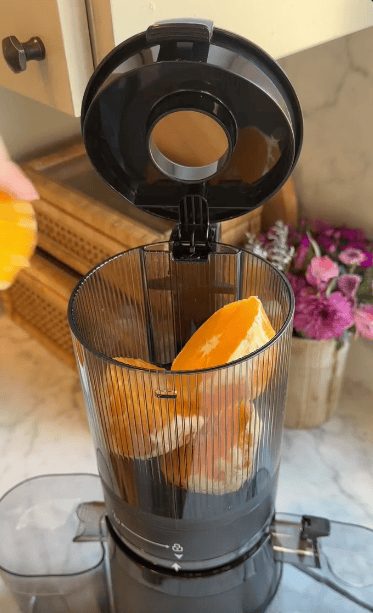 A person places peeled orange segments into the open top of a kitchen appliance juicer on a counter space, while baskets and a vase of purple flowers decorate the background.